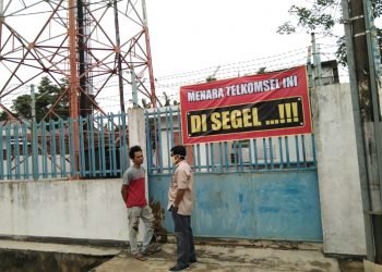 PASANG SPANDUK. Dua warga berdiri di depan spanduk yang sudah dipasang. Spanduk bertuliskan menara Telkomsel ini disegel terpampang di gerbang menara BTS tersebut. n LAMPUNG POST/BAMBANG PAMUNGKAS